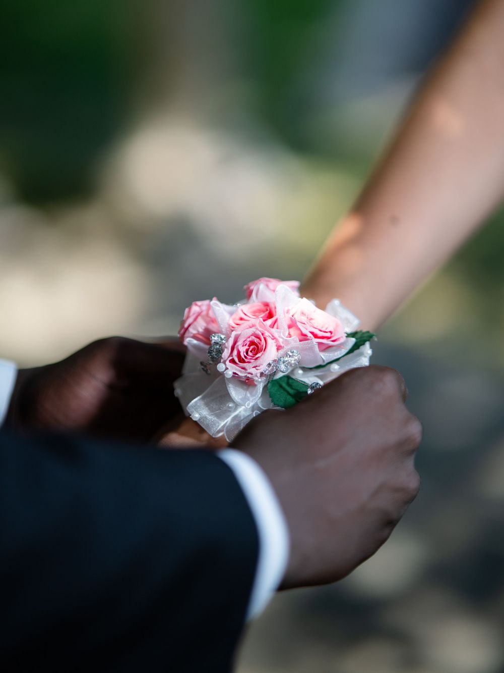 A delicate pink rose corsage being pinned on a wrist with formal attire visible in the background.