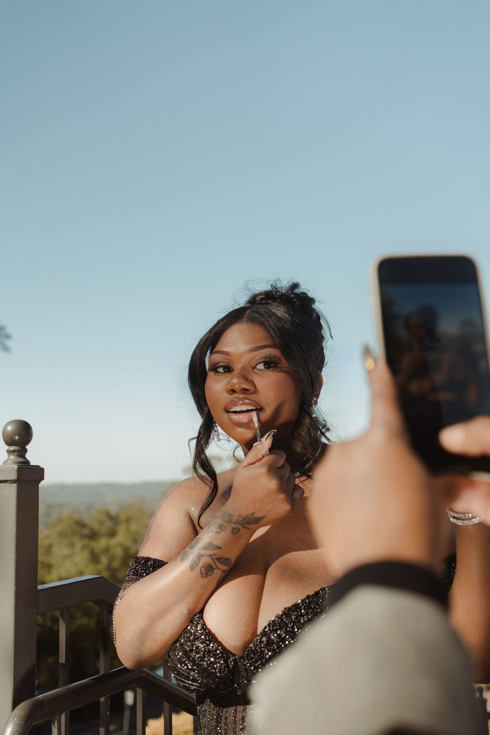 A woman in a sparkly black dress poses outdoors against a blue sky while taking selfies on a sunny day.