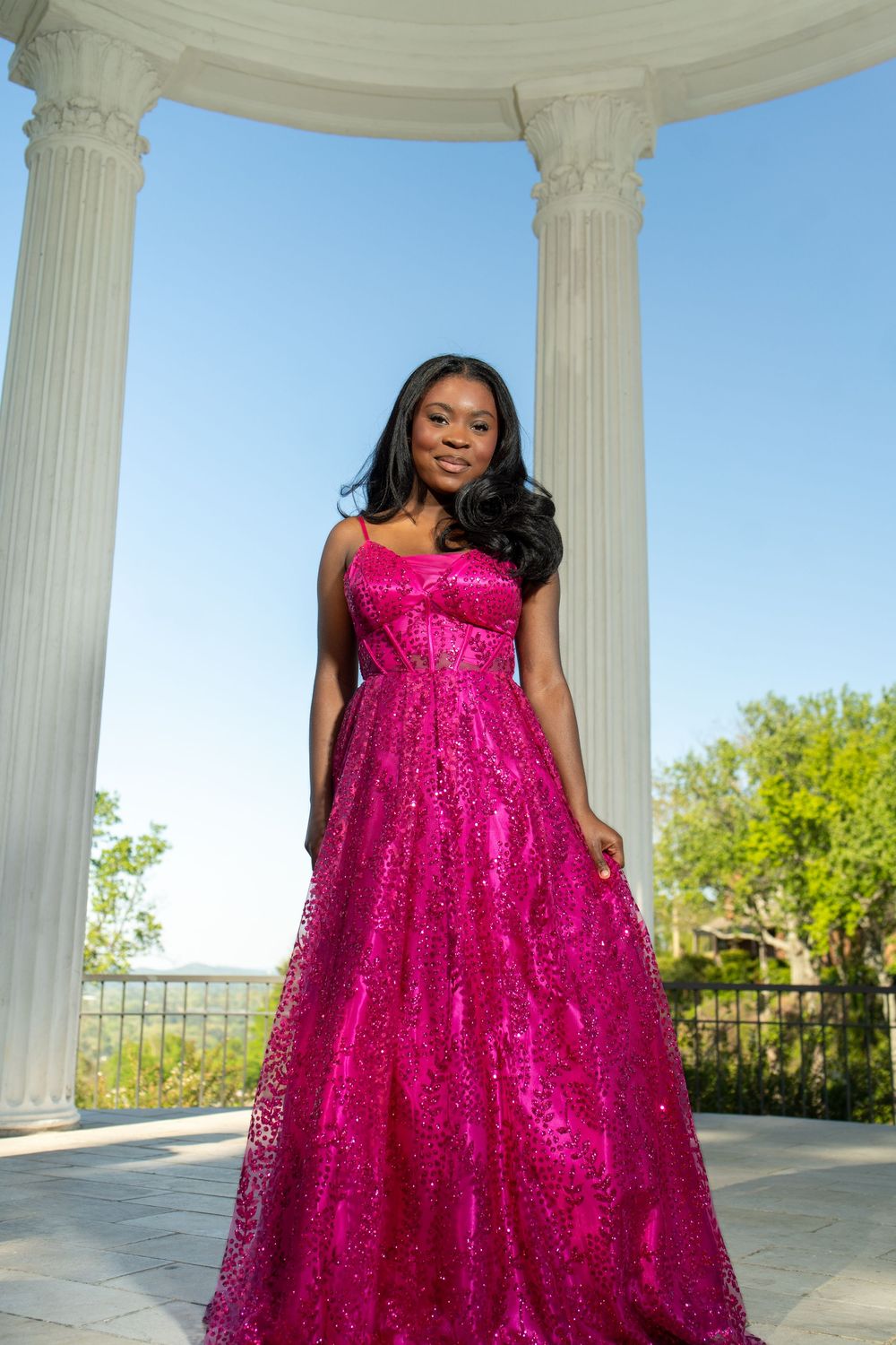 A stunning magenta sequined ball gown sparkles in the sunlight against white classical columns in an outdoor setting.
