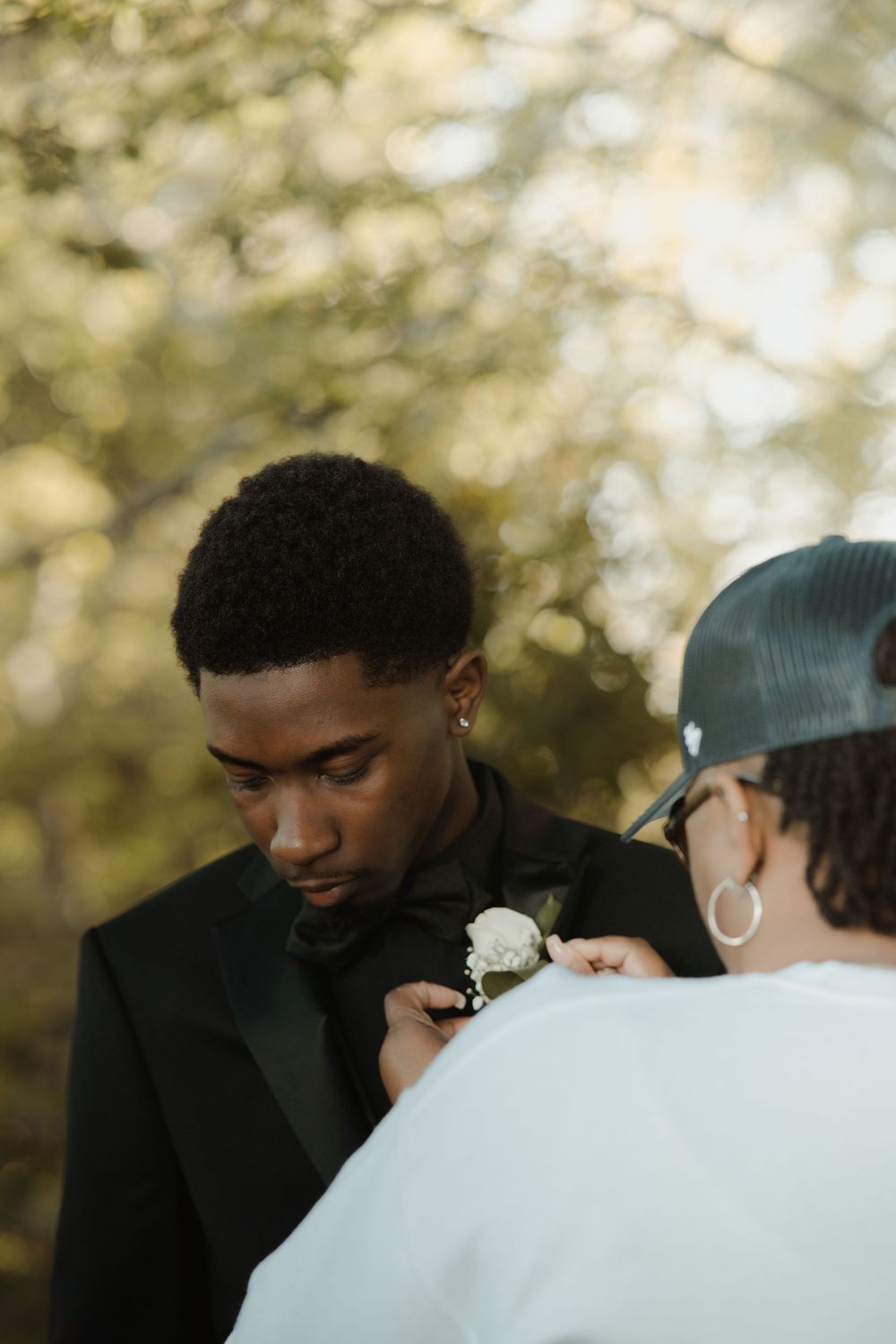 Someone helps pin a white boutonniere flower onto a black formal suit jacket at an outdoor event.