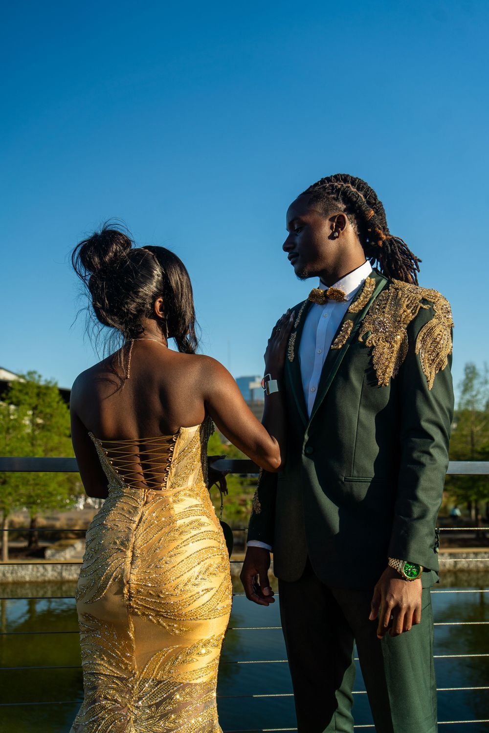 A couple in formal attire, with a gold sequined dress and dark suit, pose together by a waterfront railing at sunset.