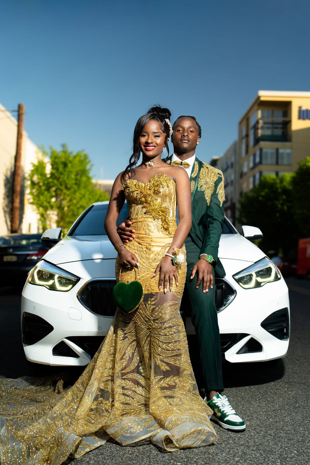 A couple in formal attire poses in front of a white BMW car in an outdoor parking lot at sunset.