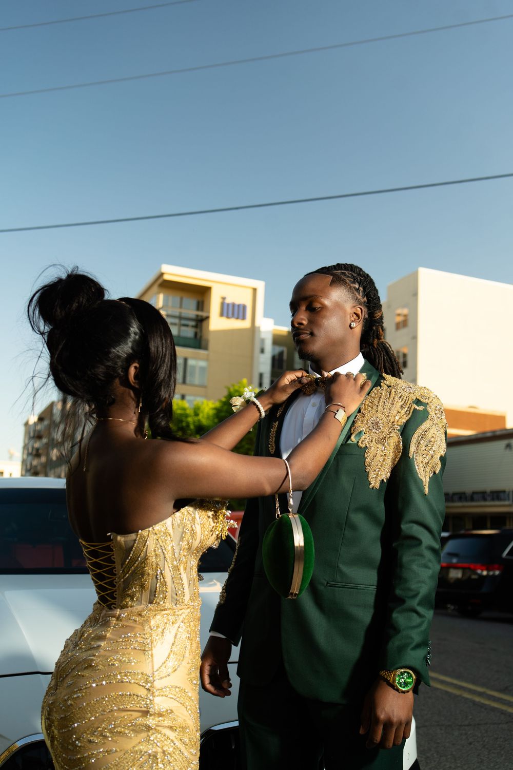 Two people in contrasting outfits, one in a gold dress and one in green, share a drink outdoors at sunset.