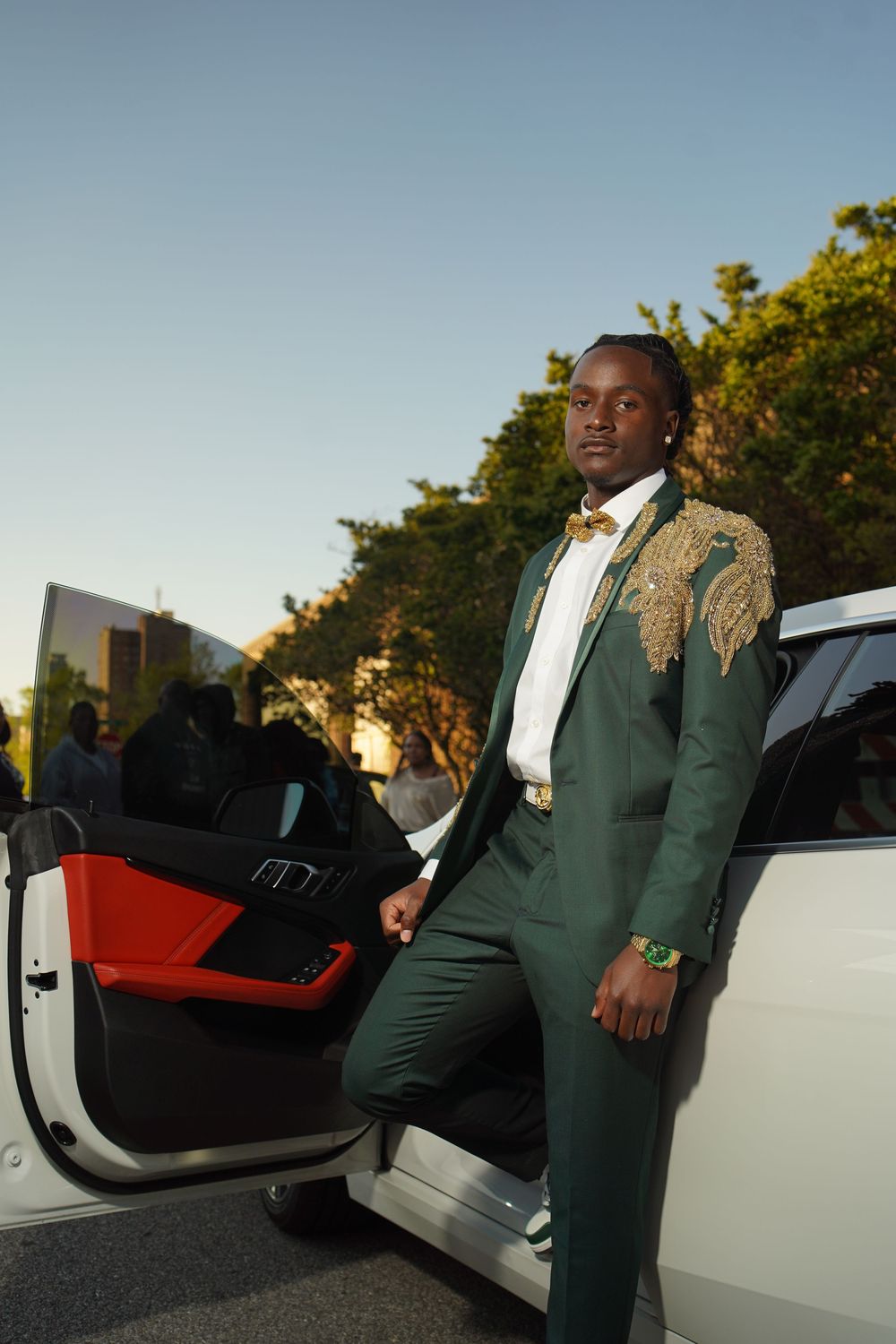 A person in an elegant green and gold embroidered suit steps out of a white luxury car at sunset.