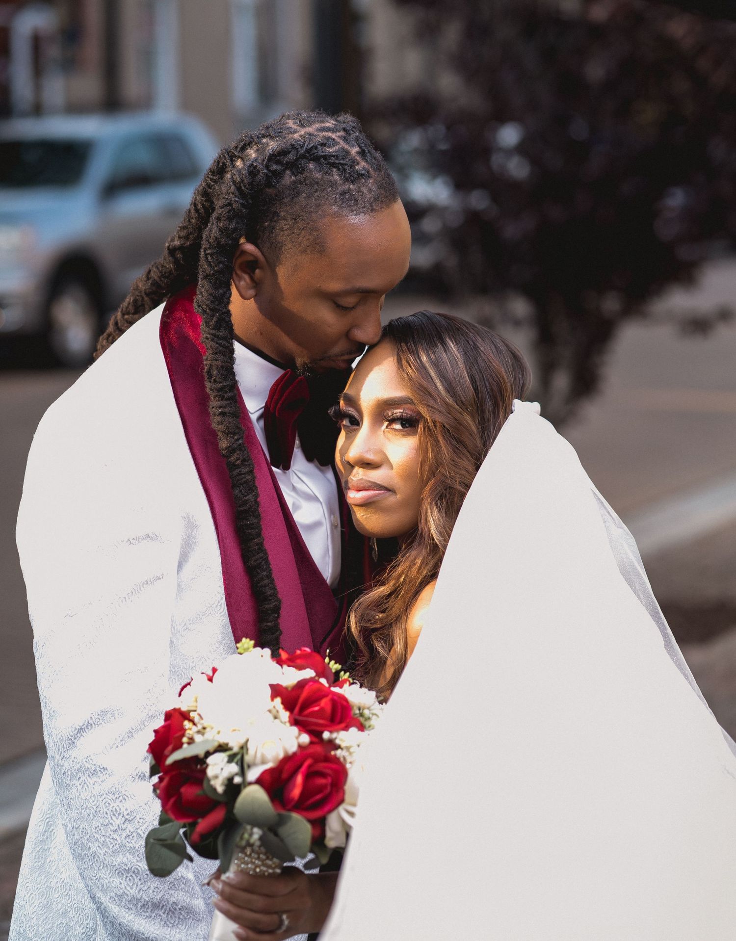 A romantic wedding couple embraces while holding a bouquet of red roses against a white and burgundy color scheme.