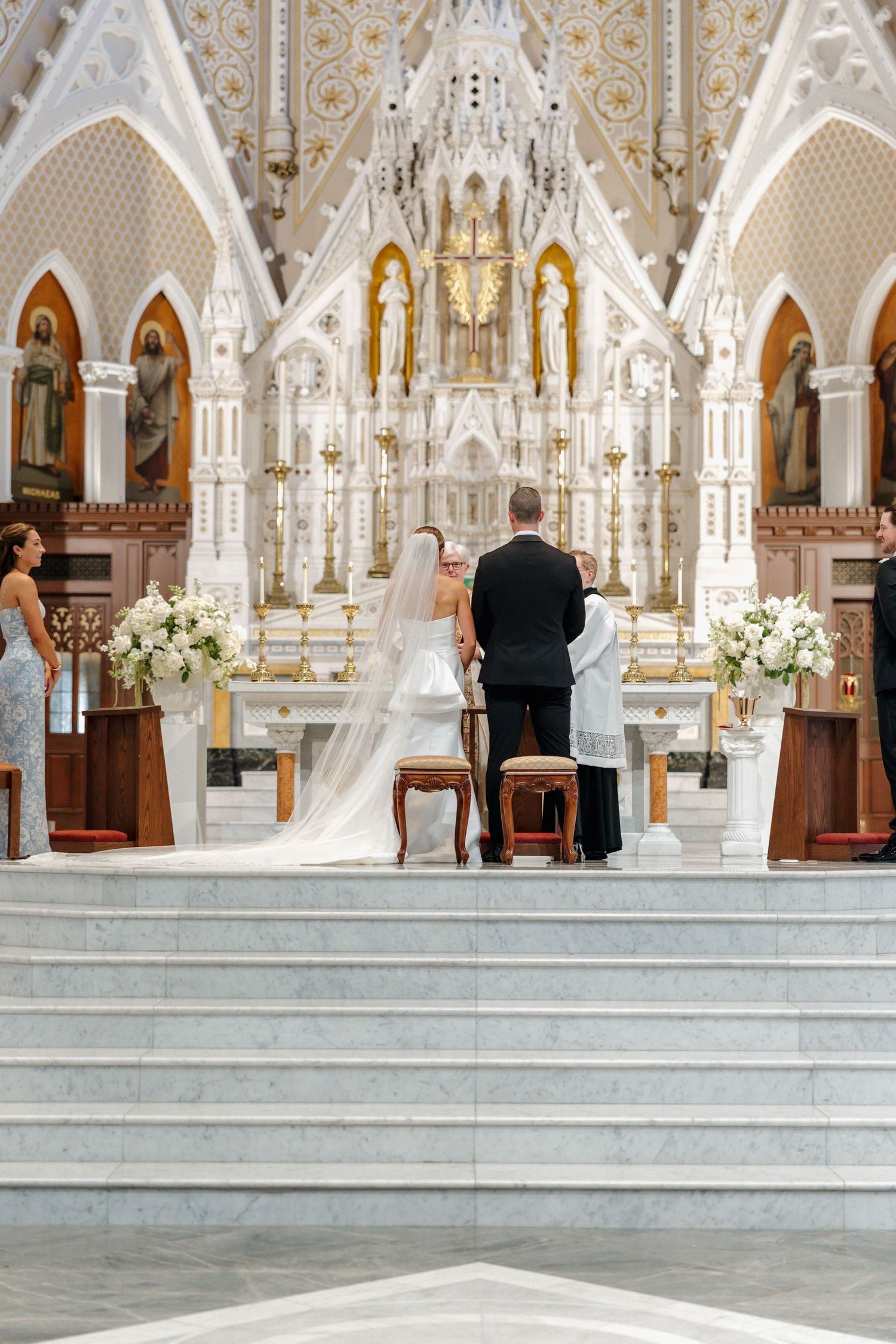 A wedding ceremony takes place in an ornate white gothic cathedral with elegant architectural details and floral arrangements.