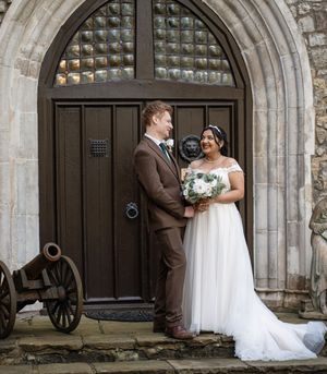 A couple in wedding attire poses together against a rustic stone archway with wooden doors and a vintage cannon nearby.