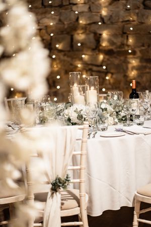 Elegant wedding reception table with white linens, candles and floral centerpieces against a stone wall with twinkle lights.