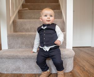 Small child sits on carpeted stairs wearing formal black vest and pants with white long sleeve shirt.