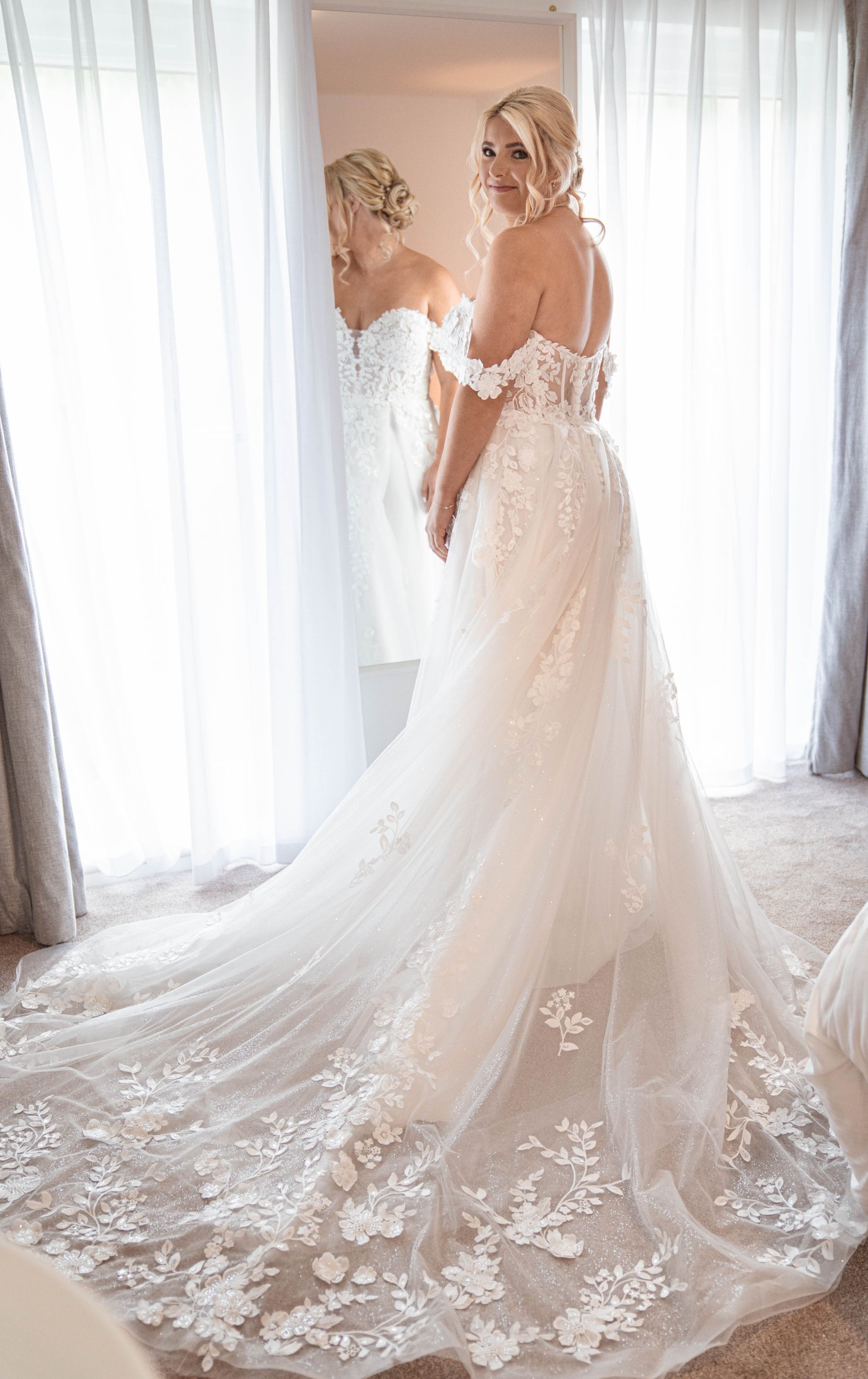 Elegant white wedding gown with intricate lace details and flowing train displayed in a bright room with sheer curtains.