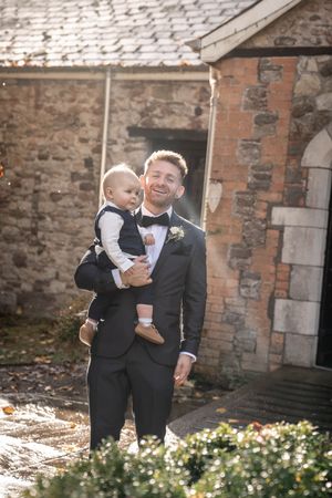 Two people in formal attire share a joyful moment together against a rustic brick wall backdrop with flowers.