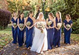 Wedding party in matching navy blue gowns stands with bride in white dress holding bouquets in autumn garden setting.
