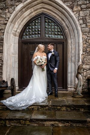 A wedding couple stands in front of a grand stone archway with a decorative wooden door at a historic church entrance.