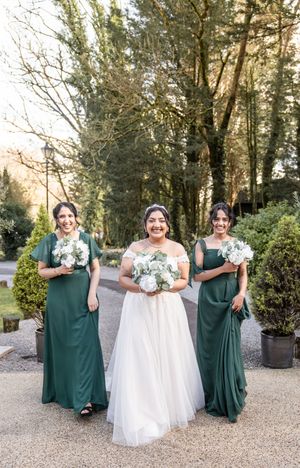 Wedding party in forest setting with bridesmaids wearing elegant emerald green dresses and bride in white gown holding bouquets.