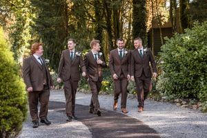 Groomsmen in brown suits walk together on a tree-lined path during a wedding photo session.