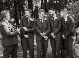 A black and white panoramic photo shows groomsmen laughing together outdoors in matching dark suits.