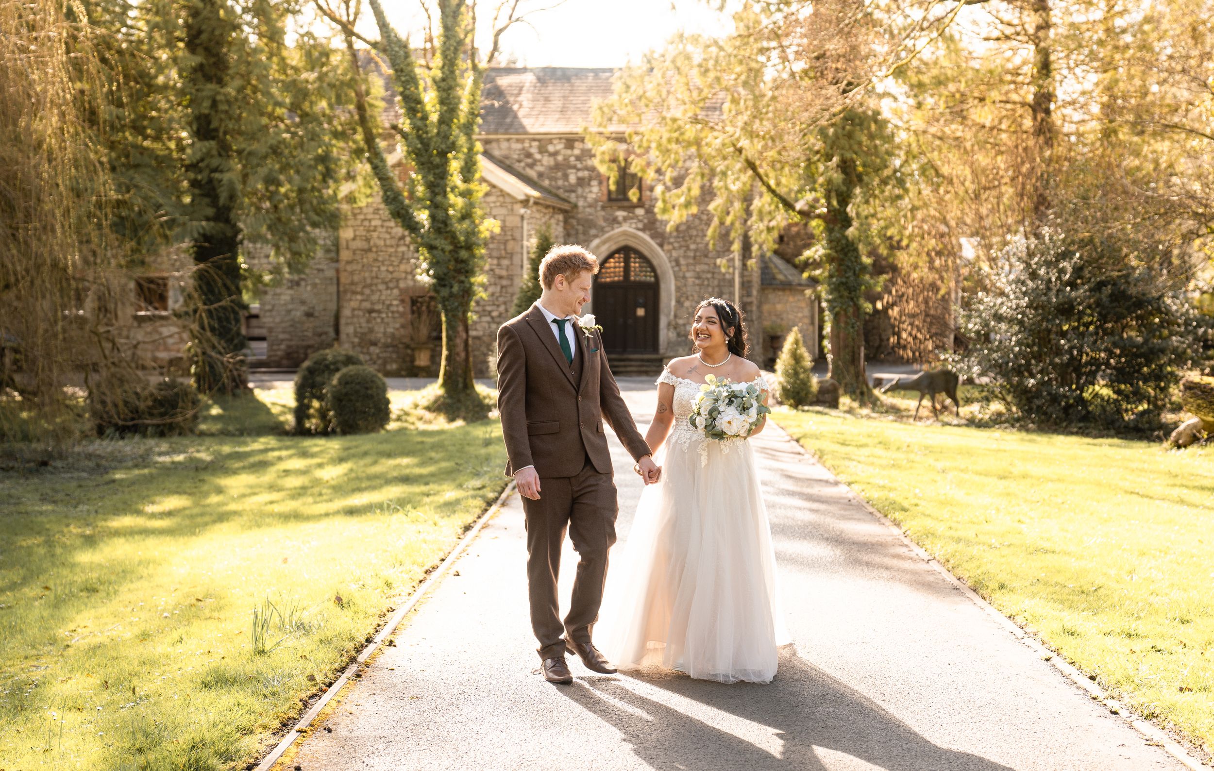 A couple walks hand in hand along a sunlit garden path beside a historic stone church on their wedding day.