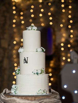 A white three-tiered wedding cake with eucalyptus accents sits on a rustic wooden base against a bokeh light background.