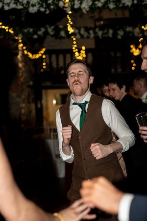 A man in a brown vest and white shirt dances energetically at an indoor party with twinkle lights in the background.