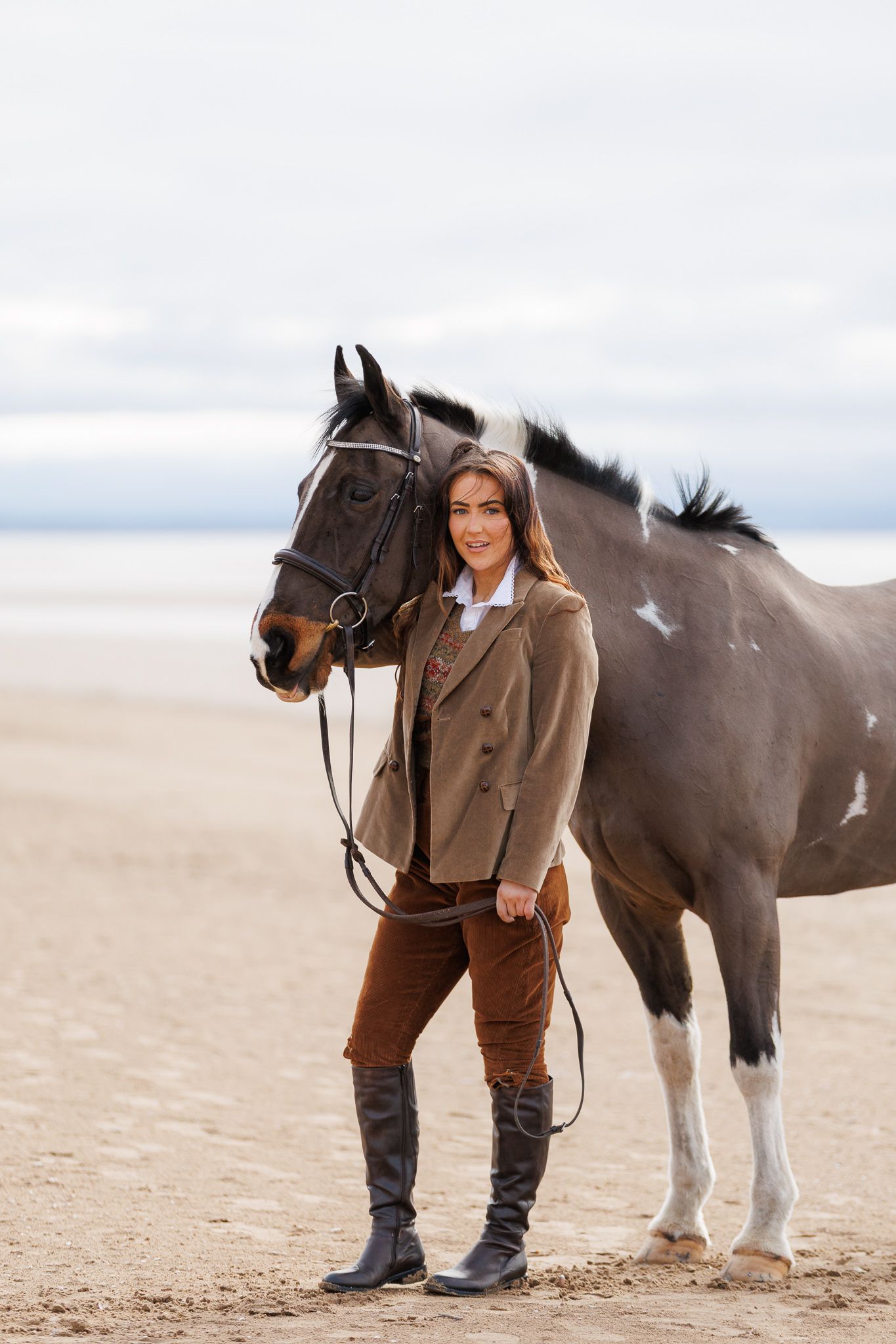 A beautiful bay horse wearing a black bridle stands next to someone in a white lace dress in an outdoor setting.