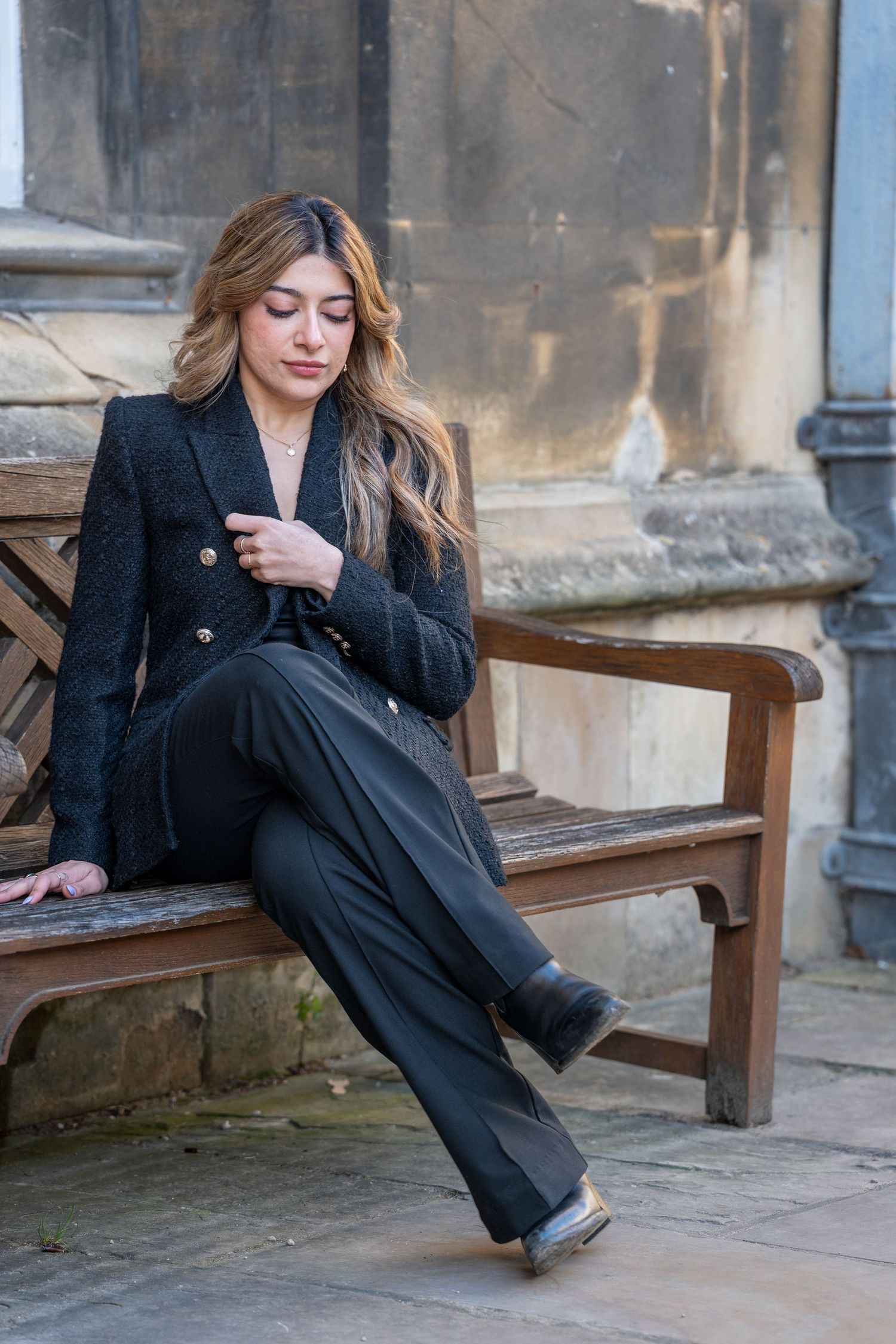 A person in a black outfit reclines on a wooden bench against a stone wall in an elegant outdoor setting.