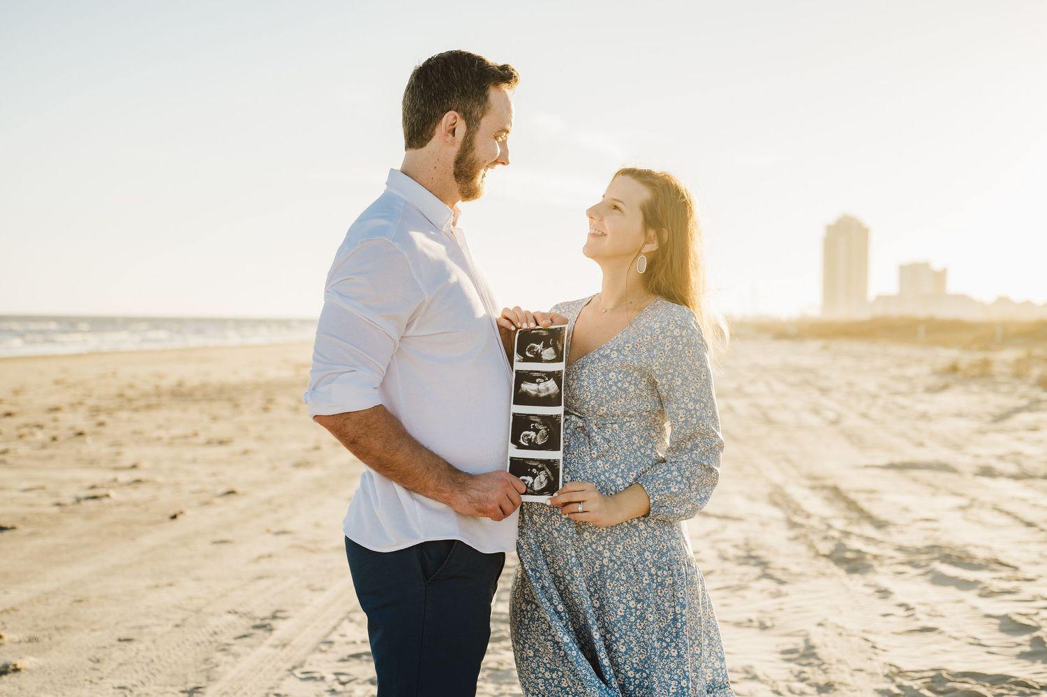 A couple stands on a beach holding ultrasound photos during a sunset announcement photoshoot.