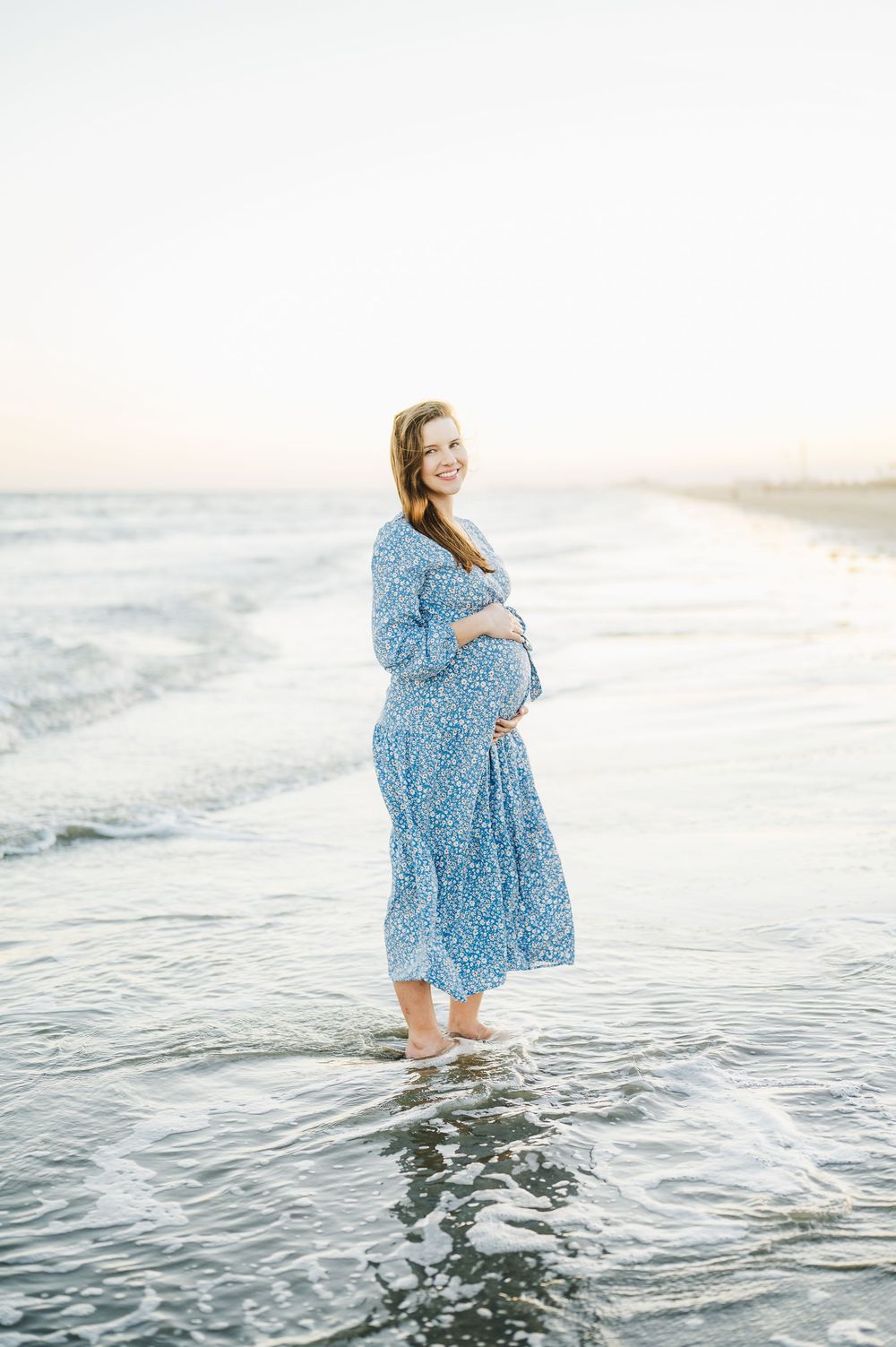 Expectant mother in flowing blue floral dress stands barefoot in ocean waves at sunset.