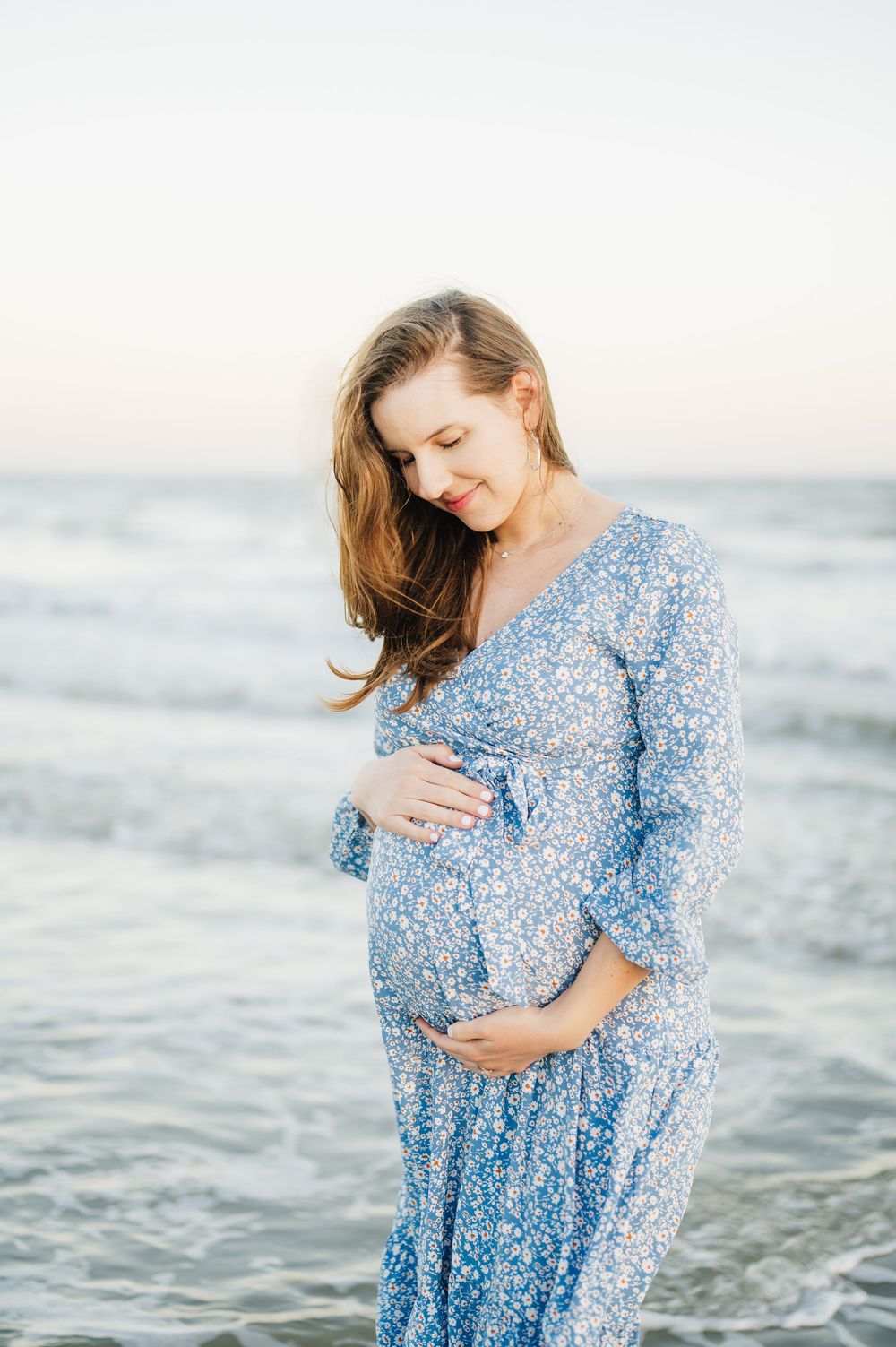 Expectant mother in flowing blue floral dress stands at ocean's edge during sunset maternity photoshoot.