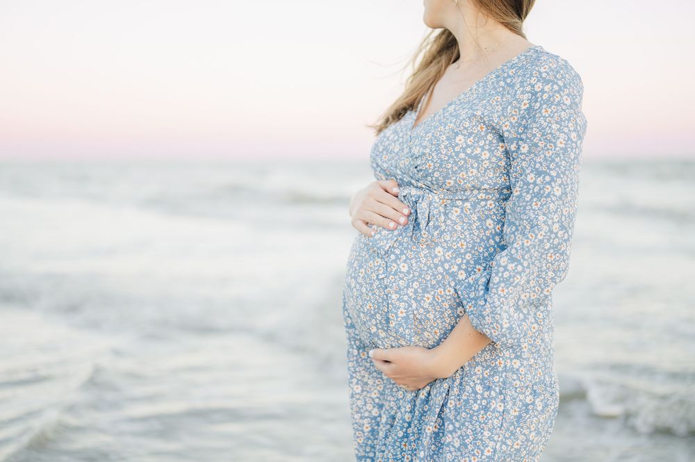 Expectant mother in a flowing blue floral dress standing by the ocean at sunset.