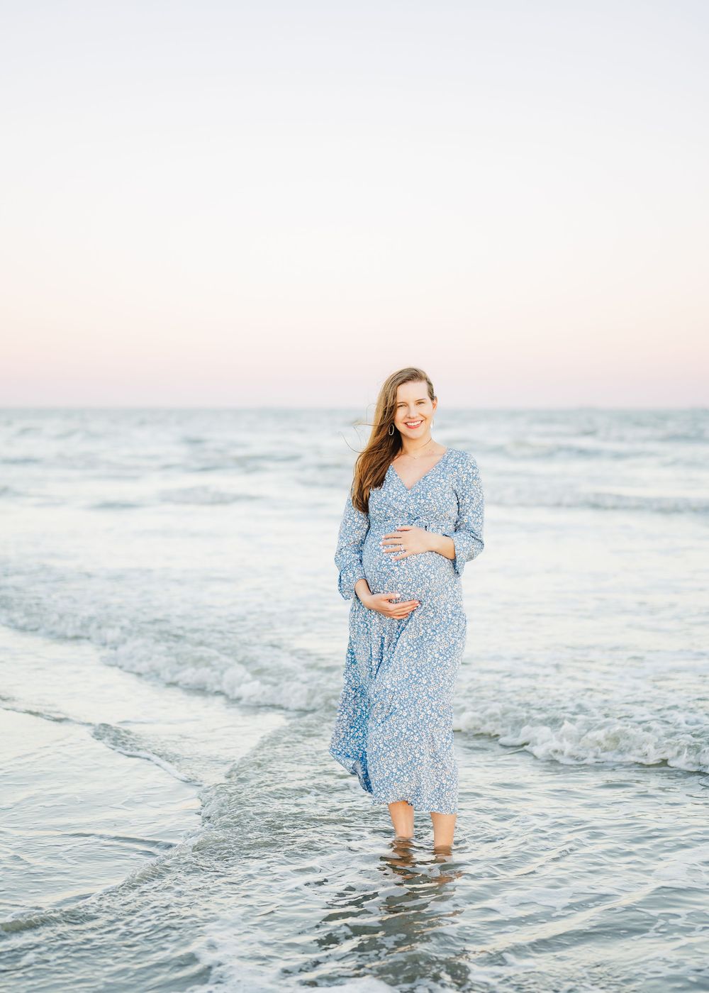 Maternity photoshoot featuring woman in blue dress walking through gentle ocean waves.