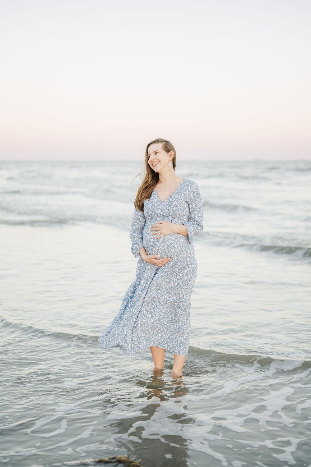 Pregnant woman wearing blue floral maxi dress stands ankle-deep in gentle ocean waves at dusk.