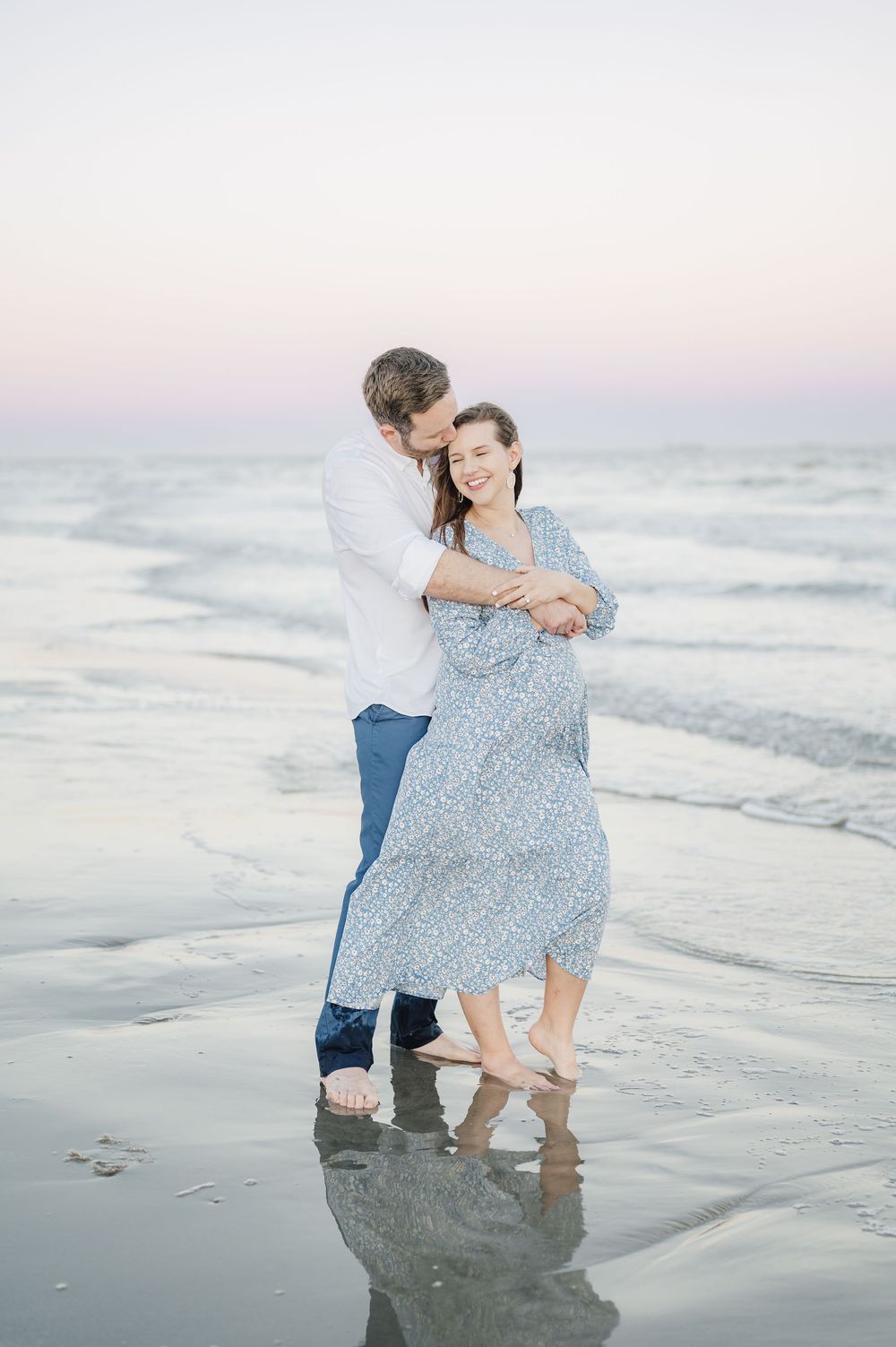 A couple embraces on a misty beach at sunset with gentle waves lapping at their feet.