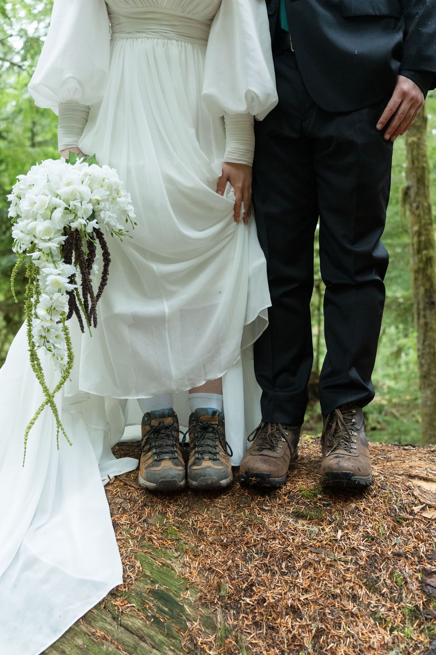 A wedding couple shows their contrasting footwear with hiking boots and a white dress in a forest setting.