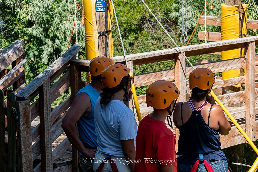 A group of people wearing orange safety helmets participate in a ropes course challenge at an outdoor adventure park.