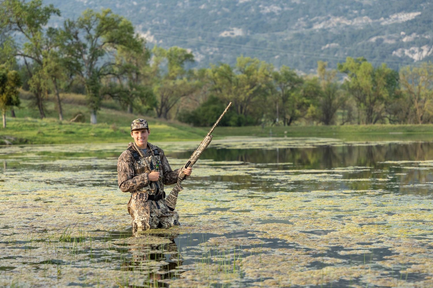 Angler wades through lily pad covered lake with fishing rod while wearing camouflage gear in mountain setting.