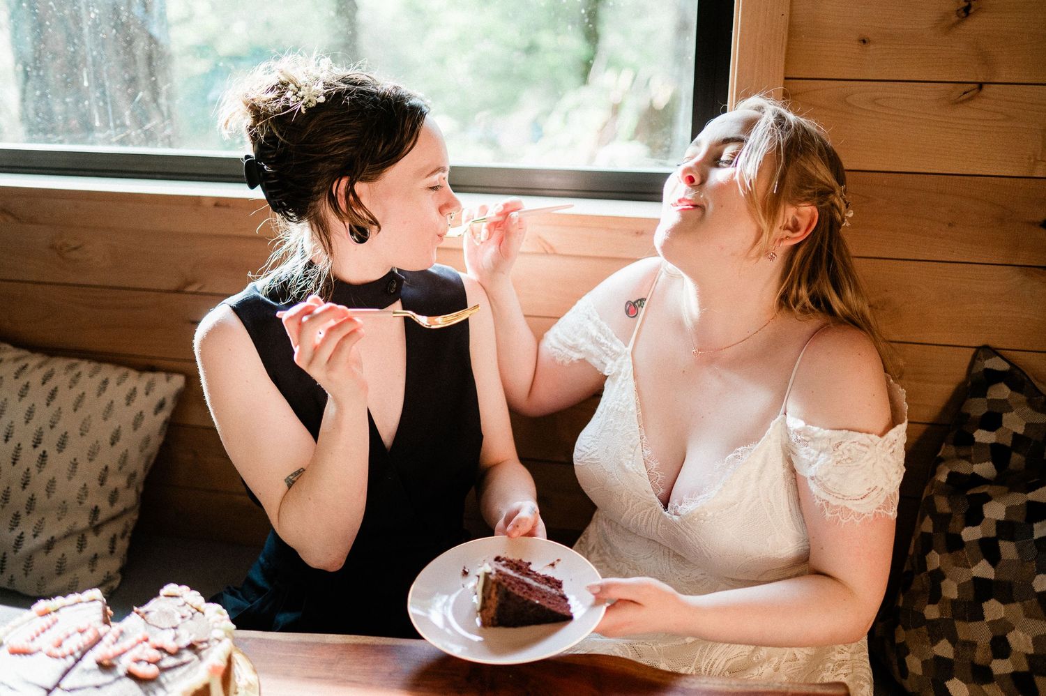 Two brides share a joyful moment while eating chocolate cake at a rustic cabin