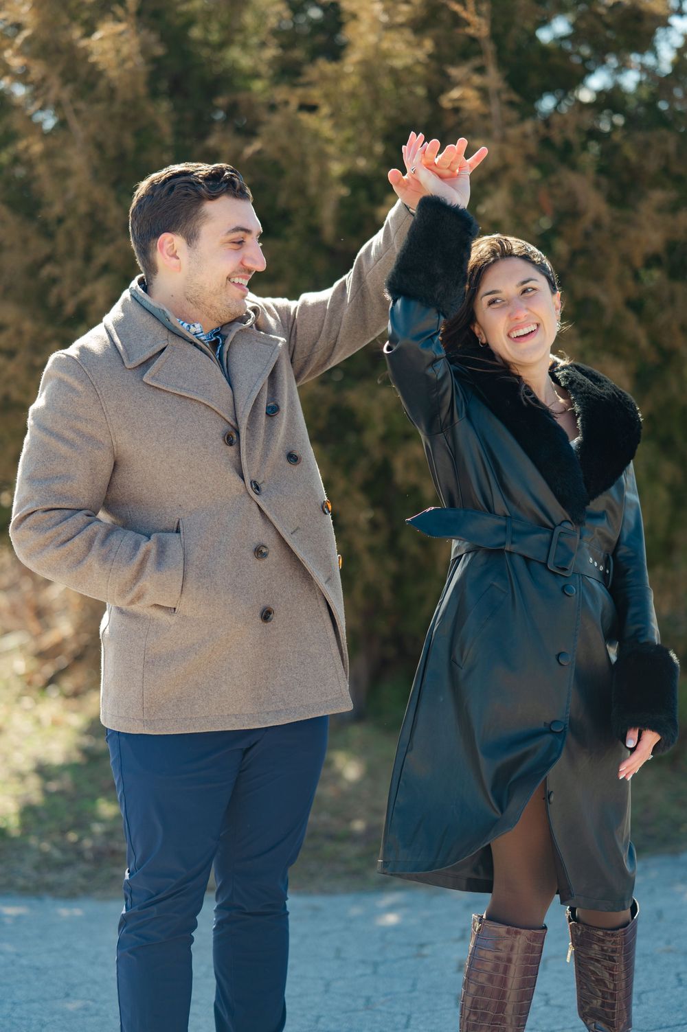 A couple in winter coats laughs and dances together on a sunny autumn day in a park setting.