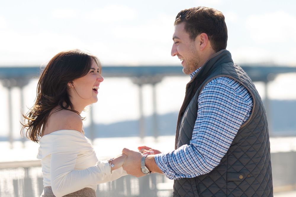 A couple embraces and laughs together during a romantic moment at a waterfront location during a proposal session.