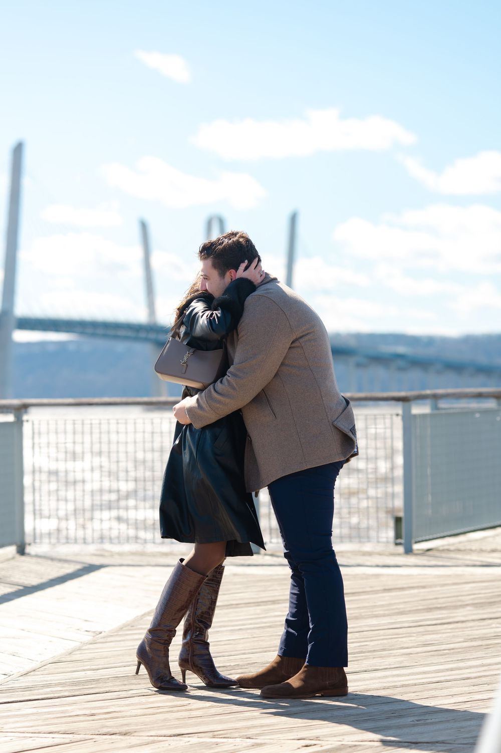 A romantic couple embraces on a wooden pier overlooking a the Hudson River on a sunny day after their proposal in Tarrytown, NY.