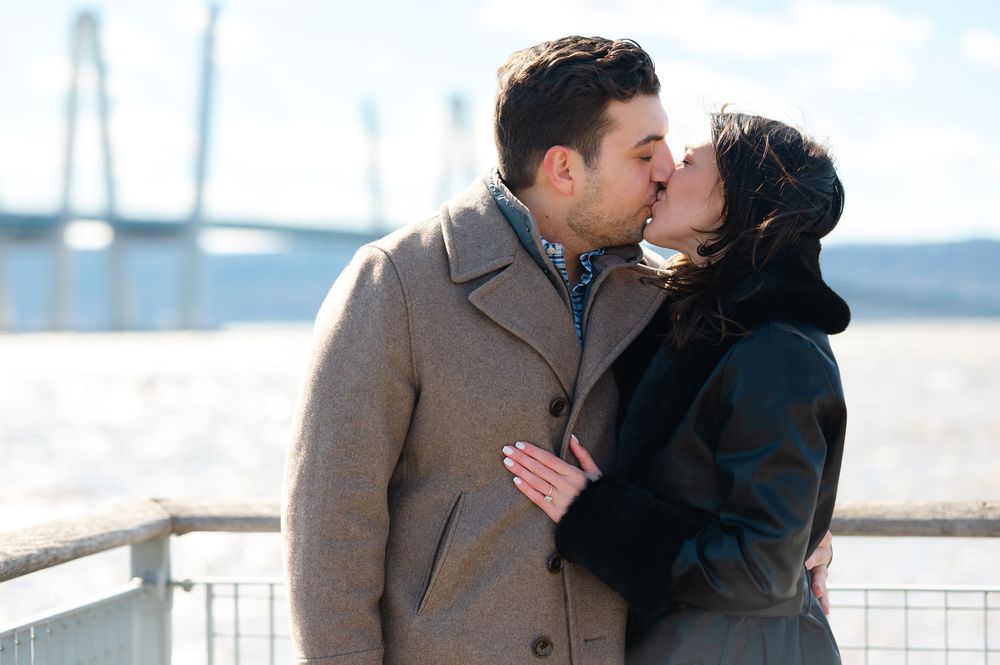 A couple shares a romantic kiss while standing on a pier with water and boats in the background.