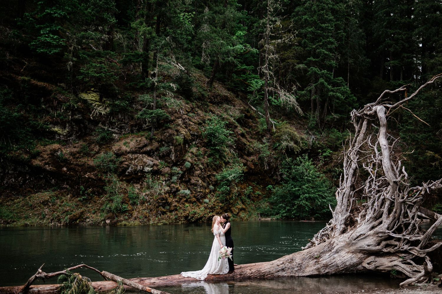 Two brides stand on a fallen driftwood tree trunk during their Packwood elopement in Washington.