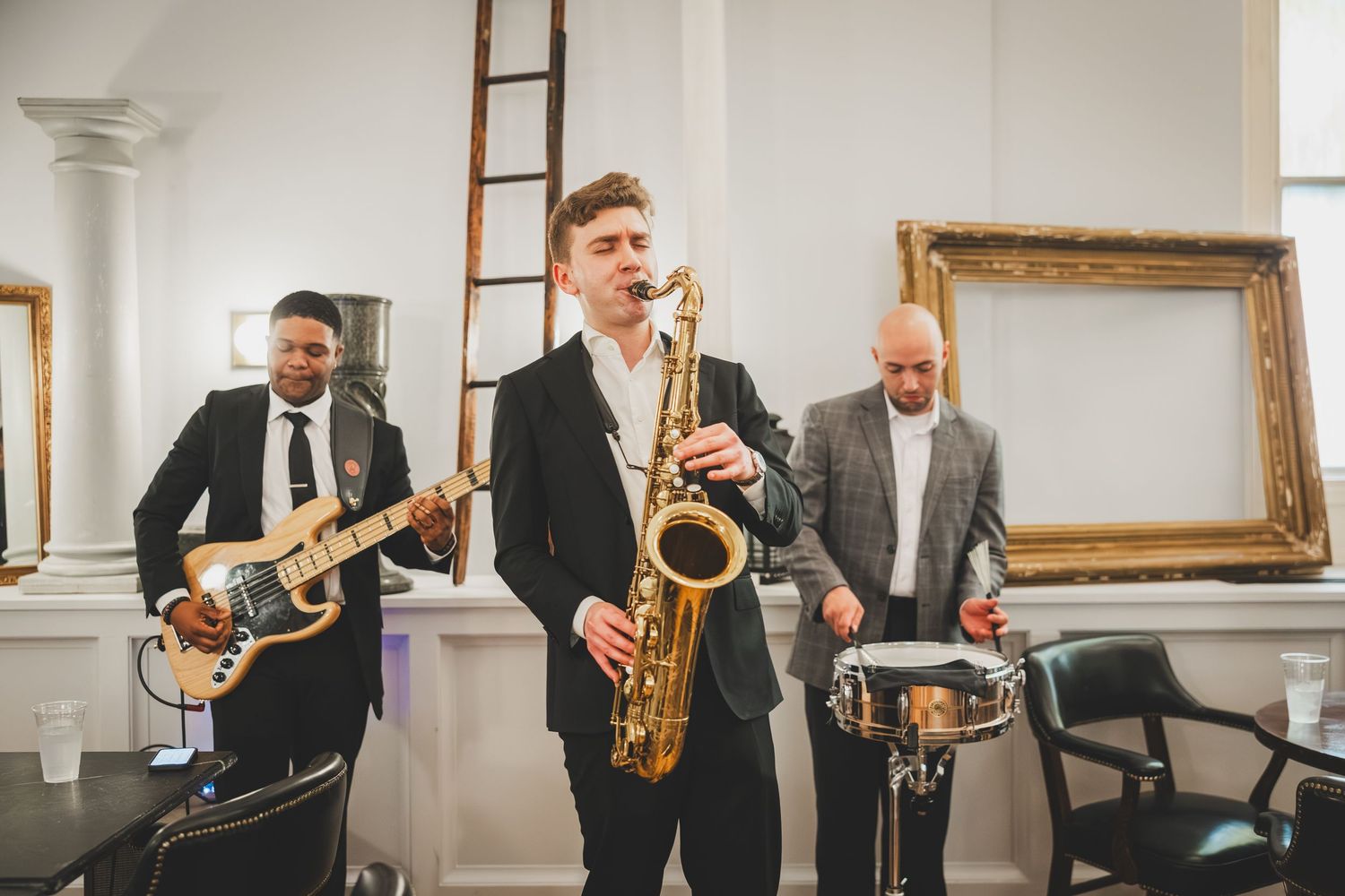 Saxophone player in black suit performs with band at indoor venue during wedding reception or event.