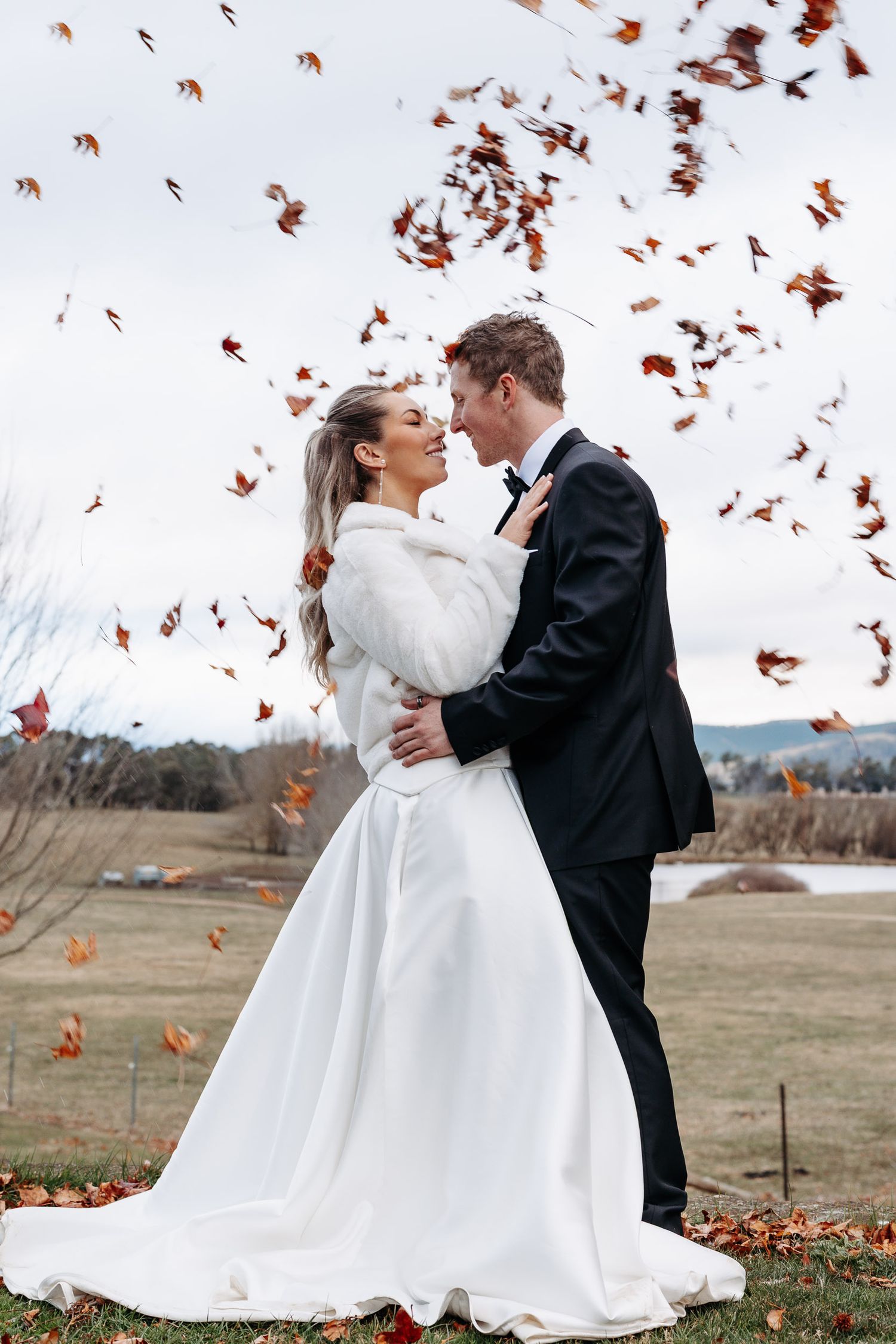 A romantic wedding couple dances together in a field as autumn leaves swirl around them in the wind.