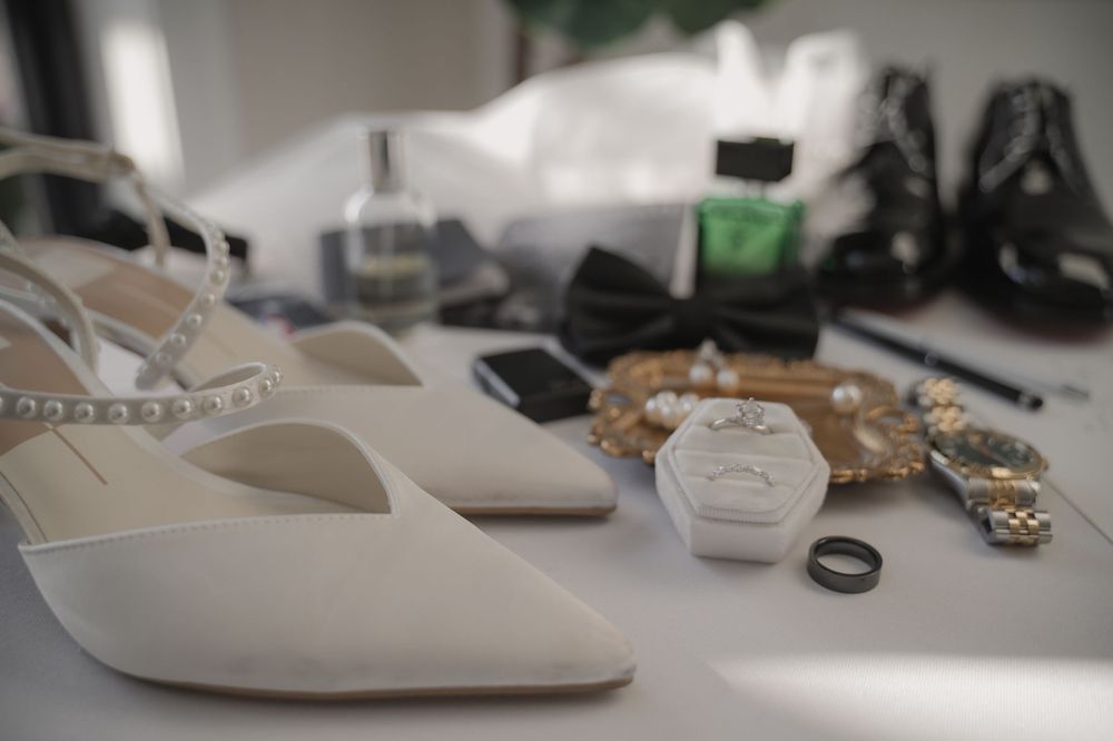 White bridal heels and wedding accessories including rings, jewelry, and a bow tie arranged on a white surface.