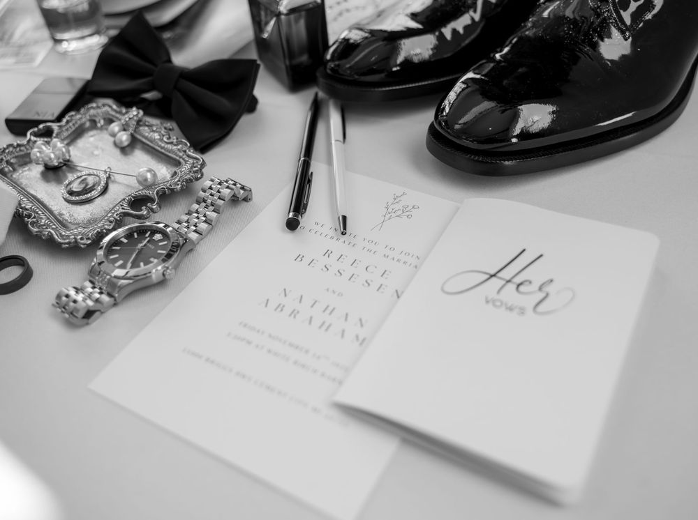 Black and white flat lay of groom wedding details including invitation, bow tie, dress shoes, and cufflinks.