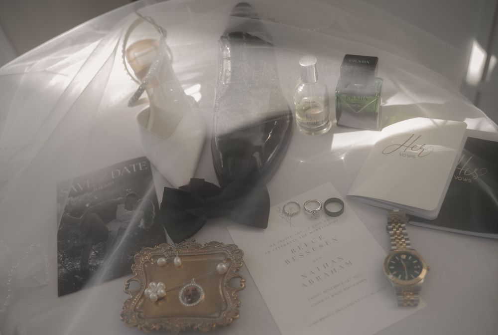 Flat lay of wedding details including rings, a watch, bow tie, heels, and accessories on white fabric.