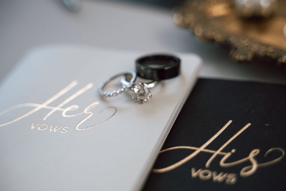 Wedding rings and vow books with gold His and Her lettering on white and black covers.