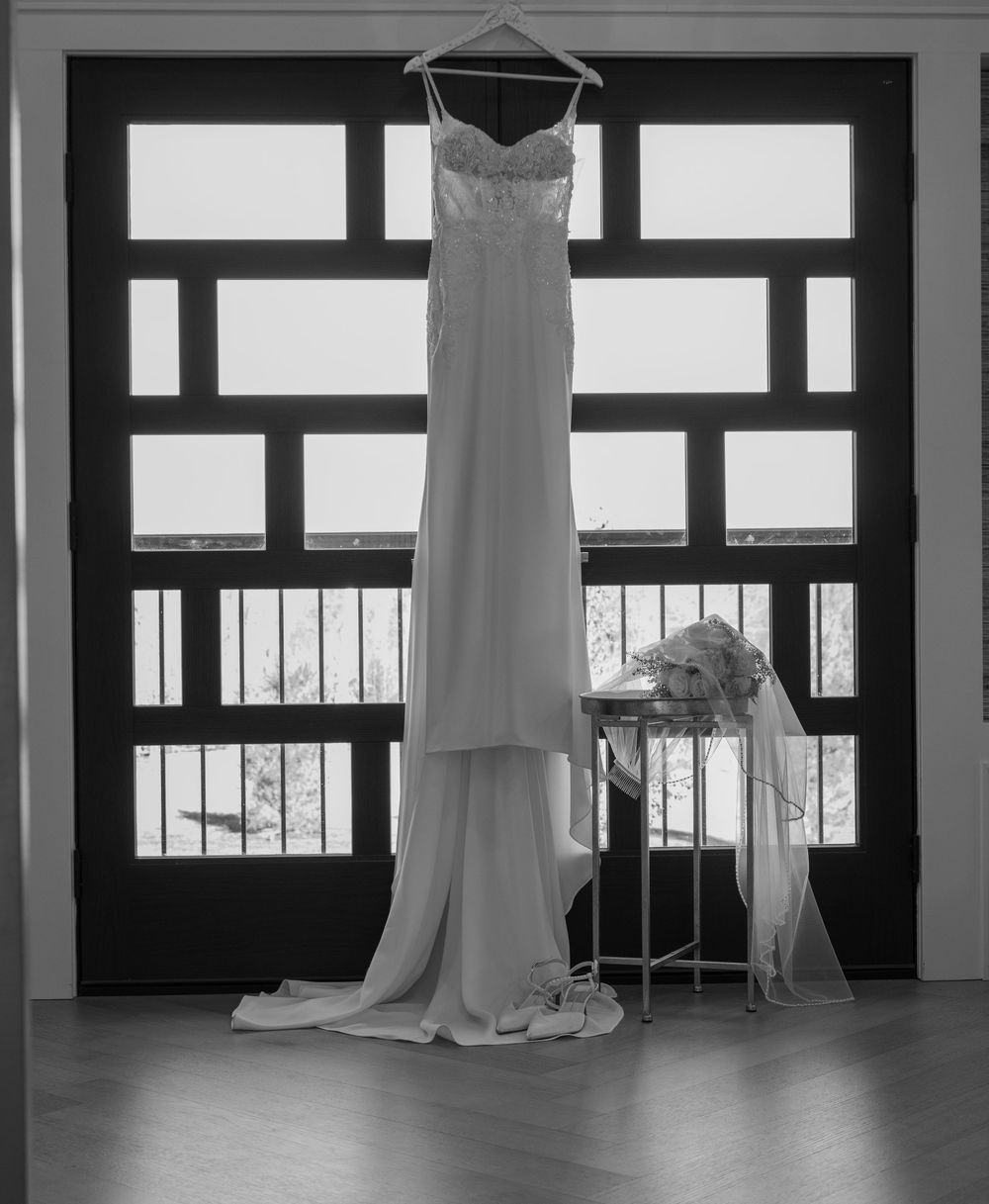 Black and white photo of an elegant wedding dress hanging by a window with a bridal bouquet on a nearby stand.