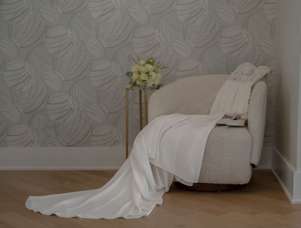 Elegant white wedding dress draped over a cream armchair beside a floral bouquet on a gold stand.