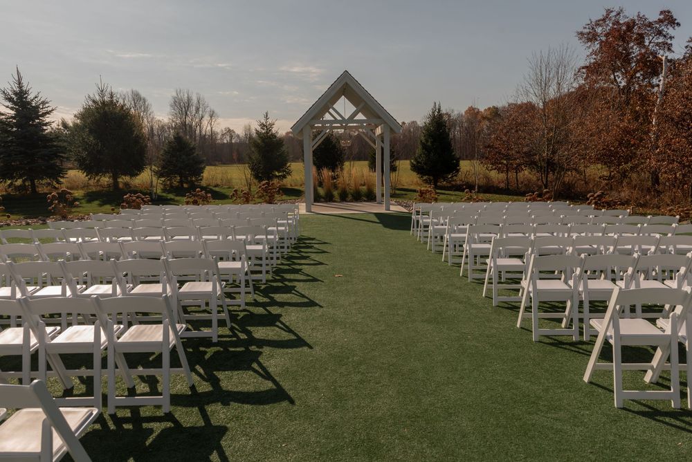 Outdoor wedding ceremony setup with white folding chairs on green grass leading to a wooden arch amid autumn trees.